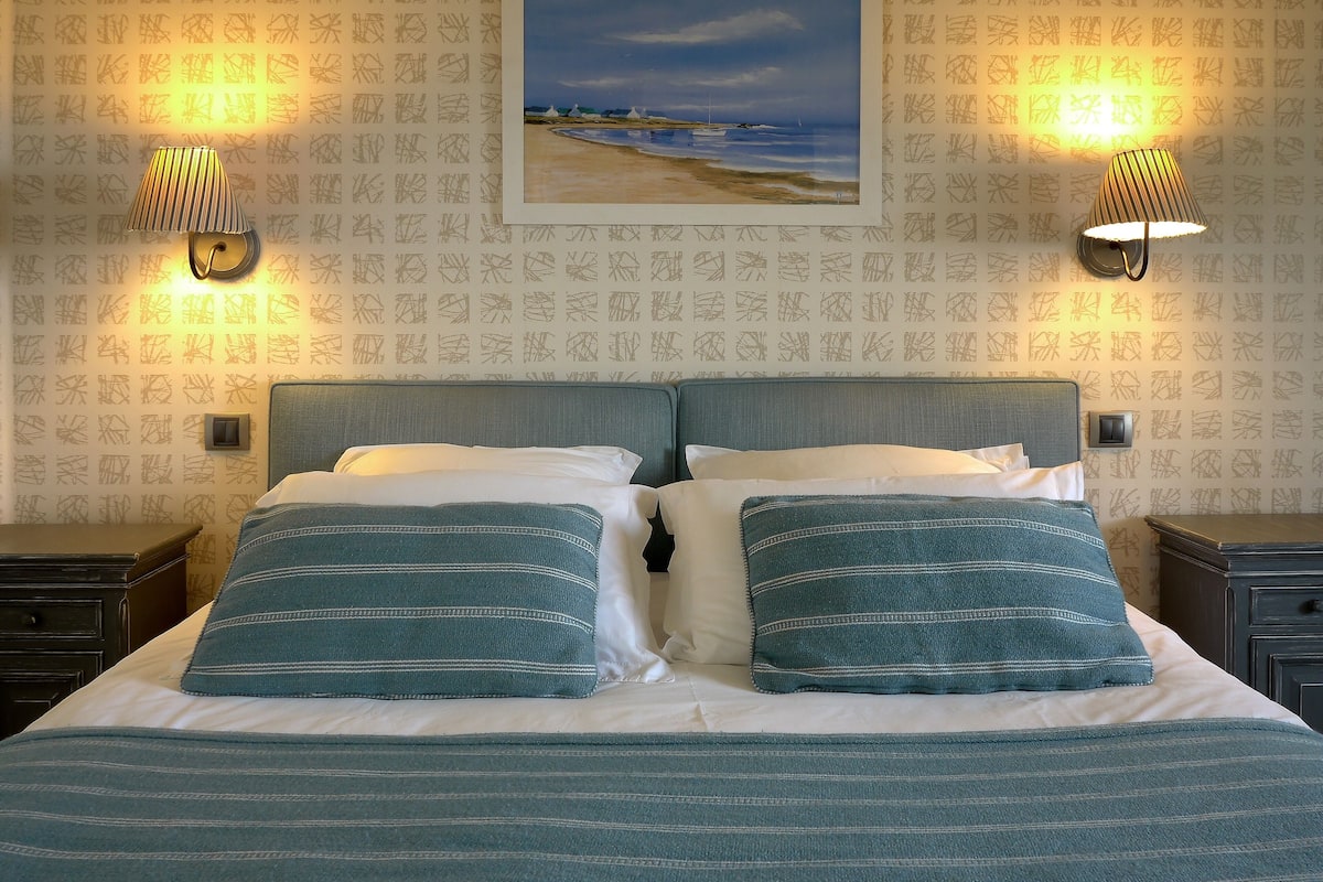 Una cama bien hecha con ropa de cama a rayas, dos lámparas de pared y un cuadro enmarcado con una escena de playa.