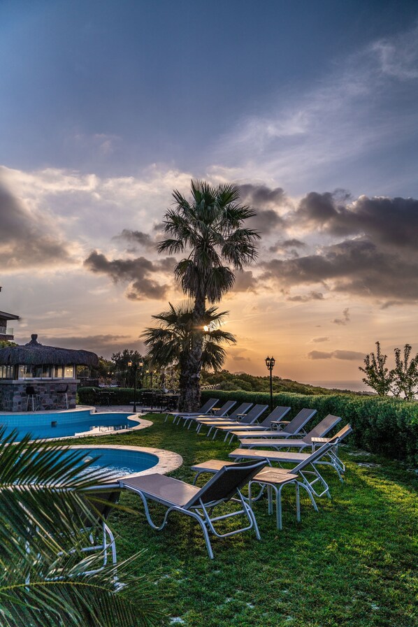 A pool area with lounge chairs and a palm tree, surrounded by greenery and a building with a thatched roof. - Assos Alis Farm Boutique Hotel & SPA (Ayvacik)