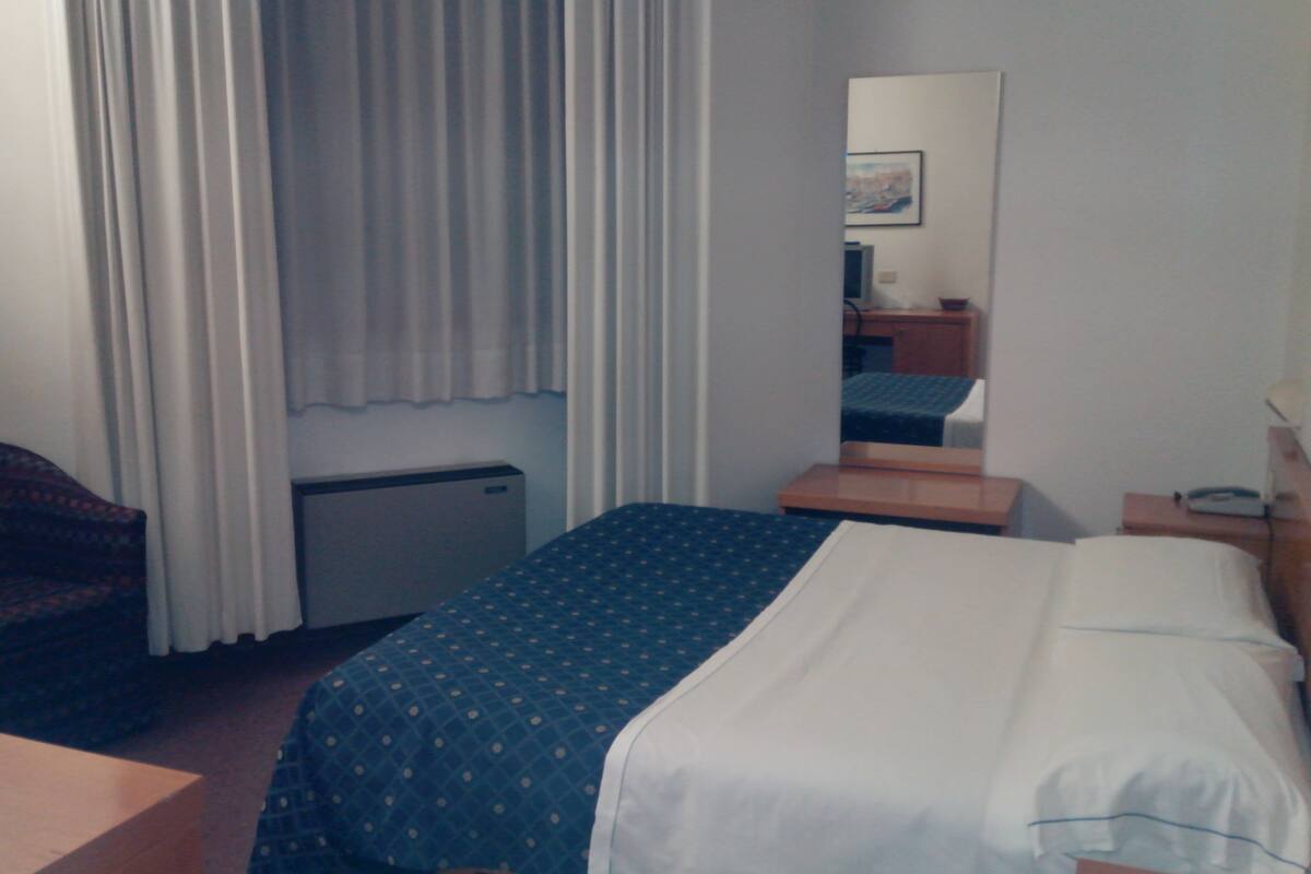 Ett hotellrum med en säng, ett skrivbord, en stol, ett litet bord, en spegel och ett fönster med gardiner.