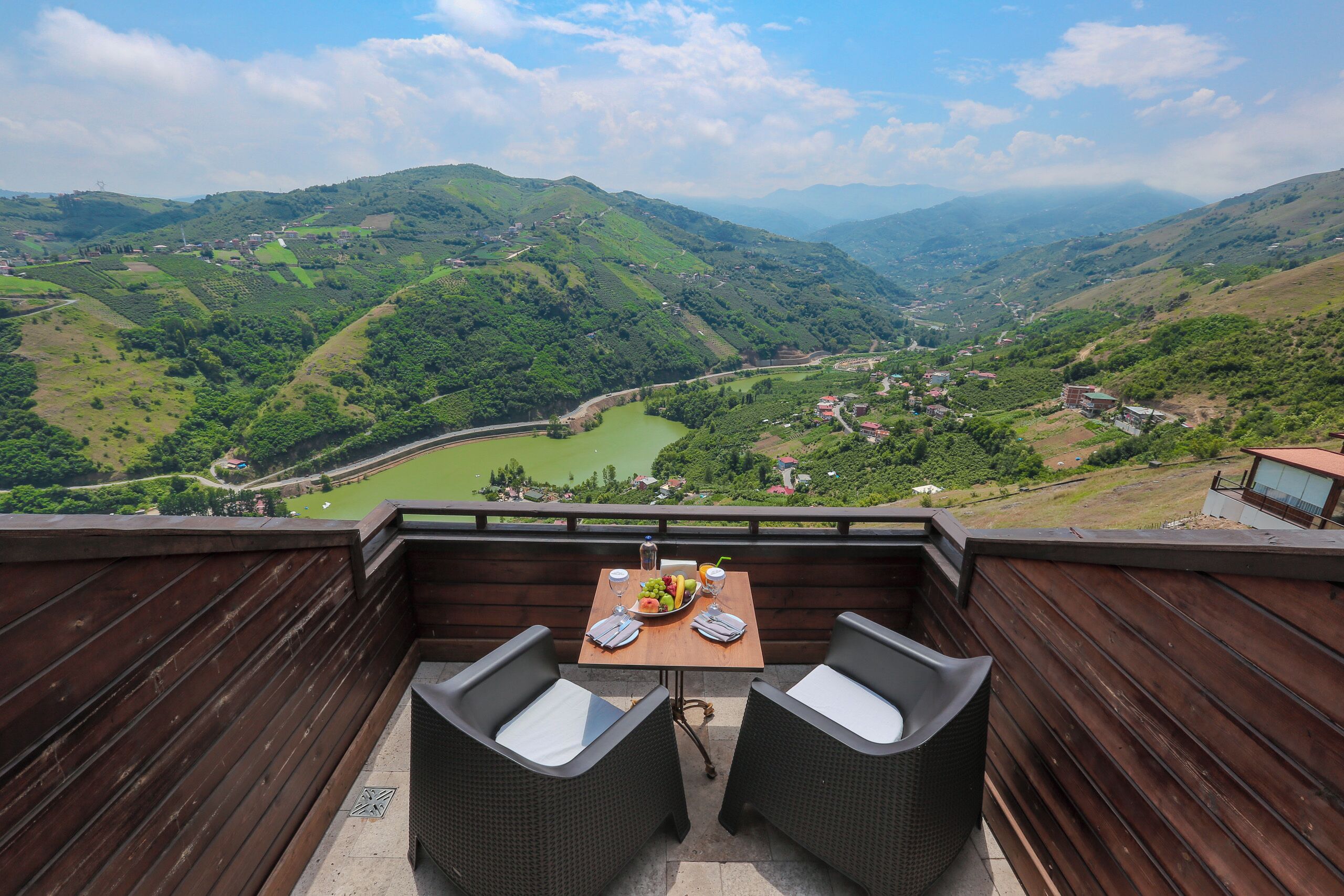 A balcony with wicker chairs and a table set for two, overlooking a valley with a river and distant hills.