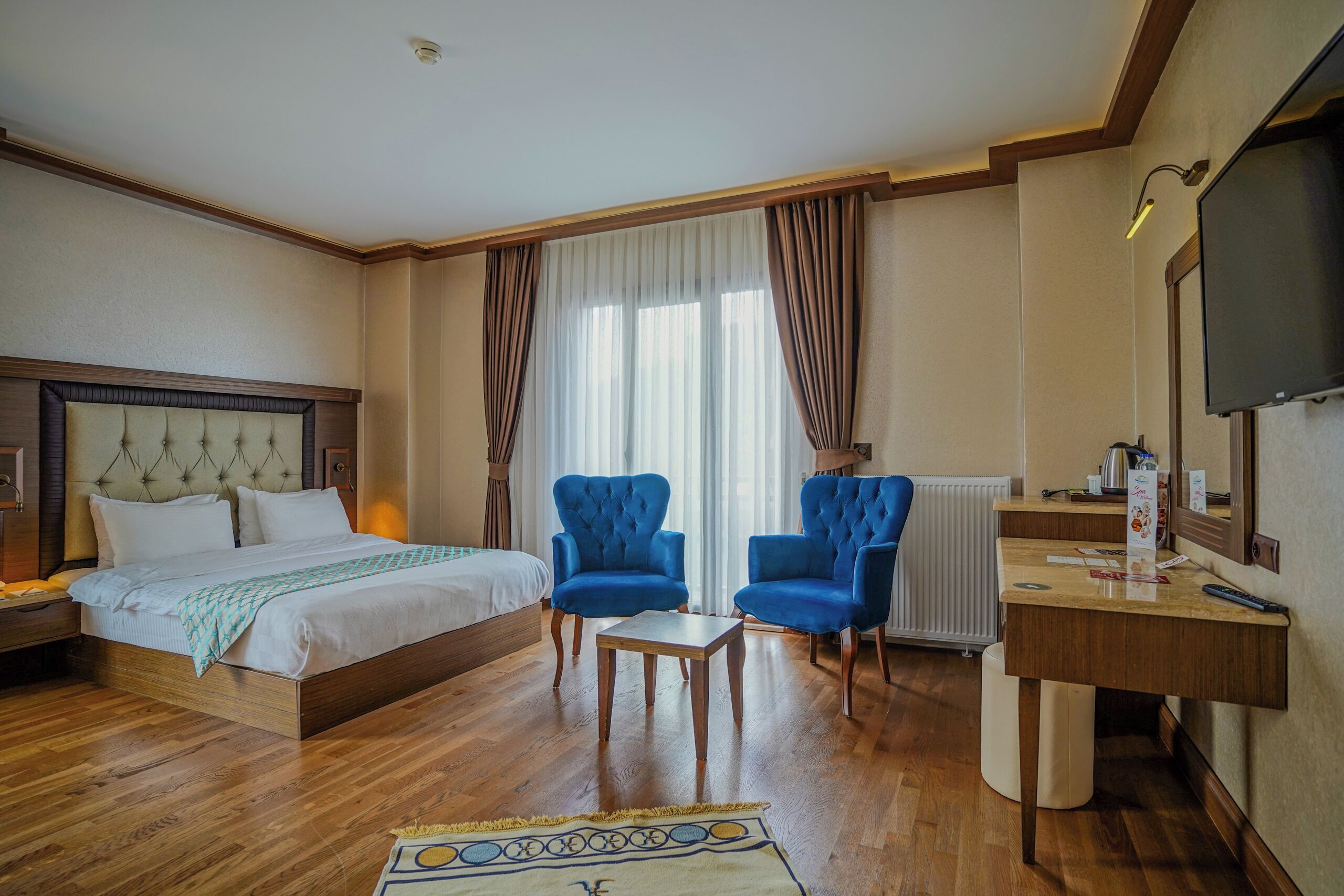Habitación de hotel con cama, dos sillones azules, una mesita, televisor y ventana con cortinas.