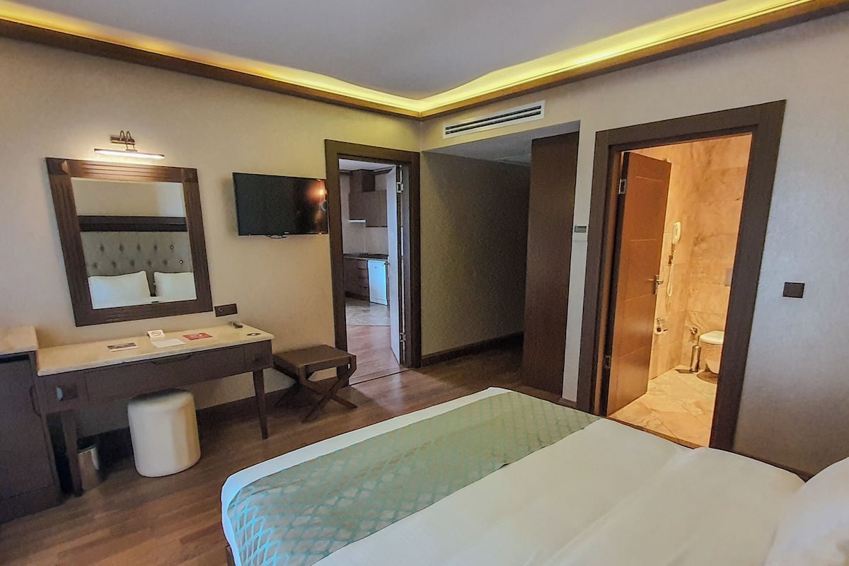 A hotel room with a bed, a bedside table, a mirror, a TV, a bathroom with a toilet and sink, and a hallway leading to other rooms.