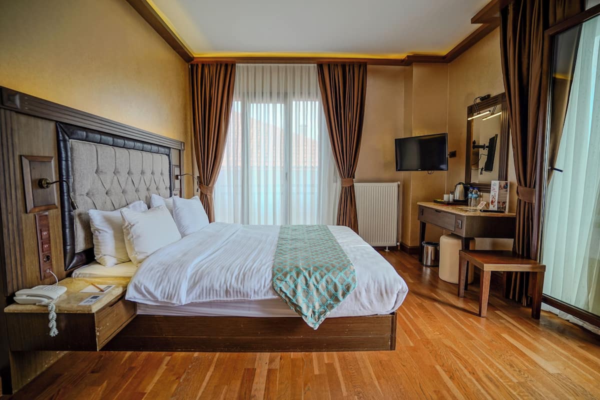 A hotel room with a bed, bedside table, lamp, TV, and a window with curtains.