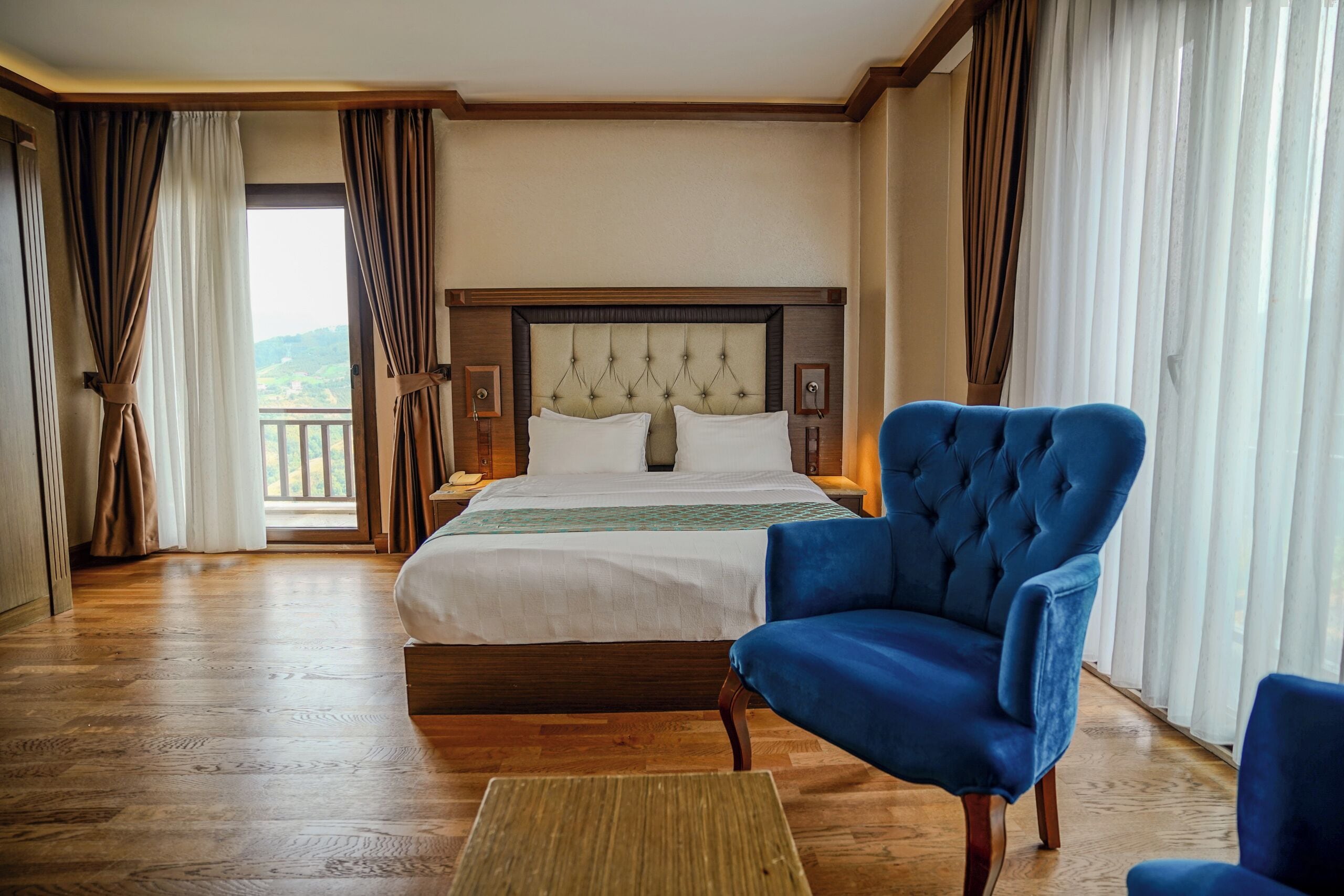 Une chambre d’hôtel avec un lit, un fauteuil bleu, un plancher en bois et une vue par la fenêtre.