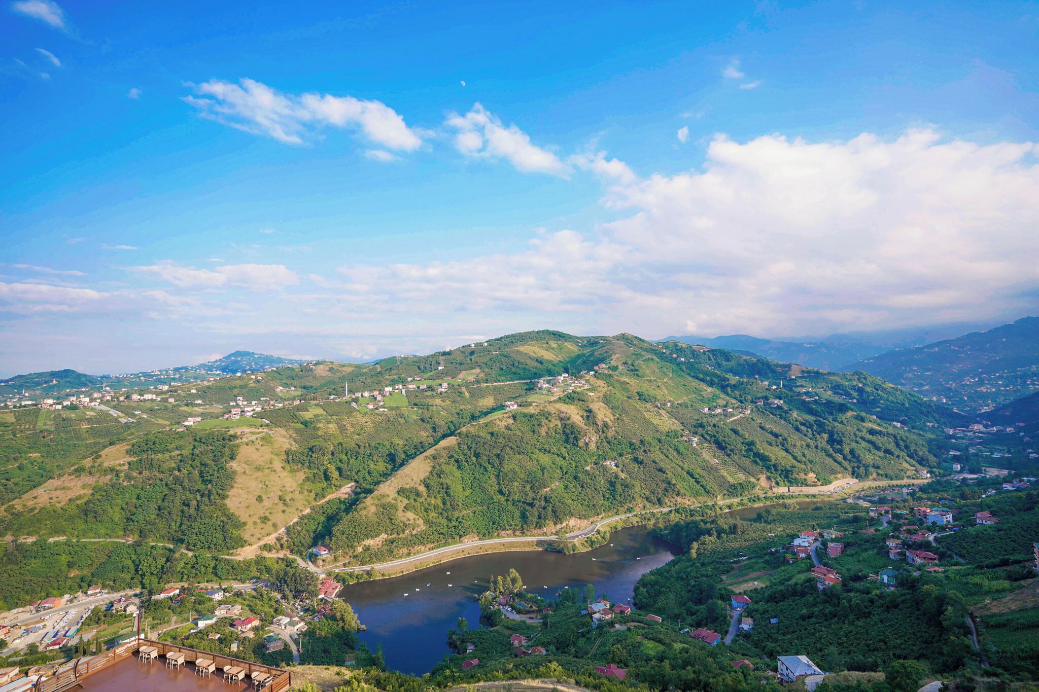 A panoramic view of a mountainous landscape with a river, a small settlement, and a wooden viewing platform.