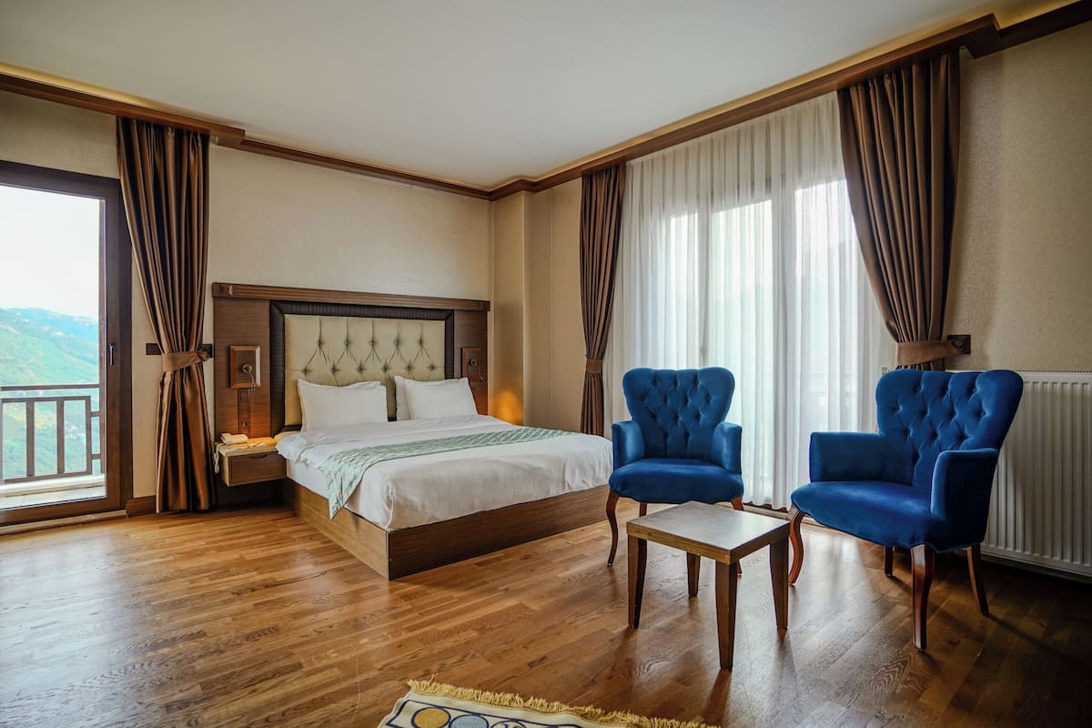 A hotel room with a bed, two blue armchairs, a small table, and a view of the outdoors.