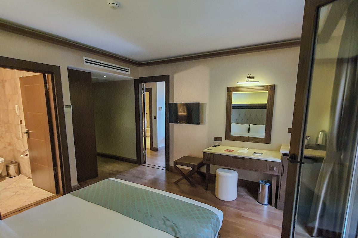 A hotel room with a bed, a desk with a mirror, a trash bin, and an air vent.