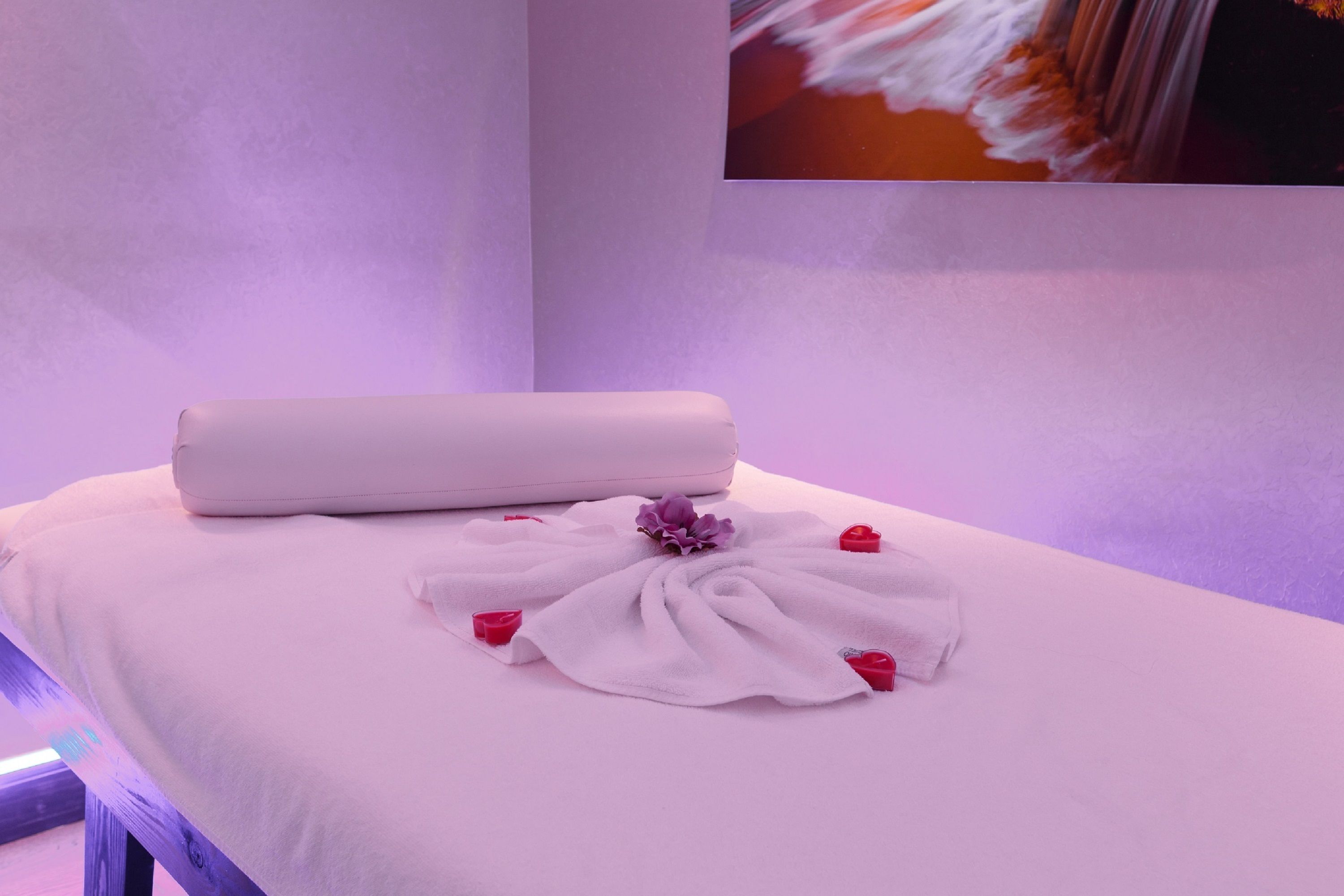 A neatly made bed with a white pillow, a white towel, and a purple flower.