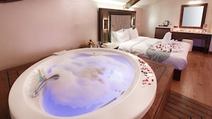 Quarto de hotel com uma banheira oval grande cheia de bolhas, piso de madeira, cama com roupa branca e cômoda de madeira com espelho.