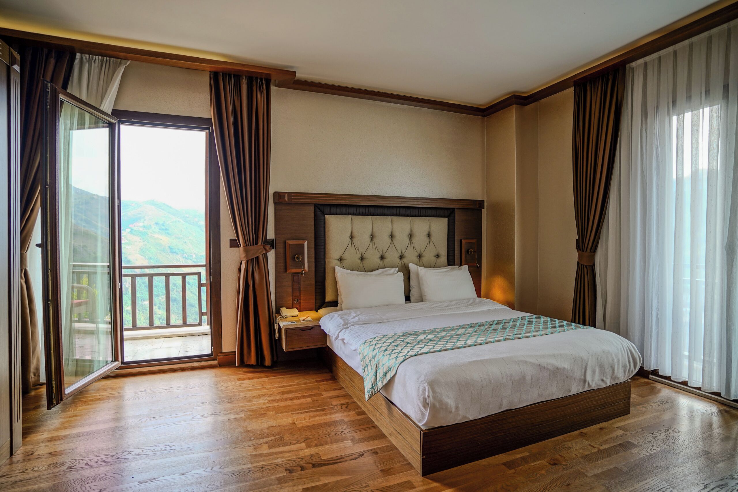 Ein Hotelzimmer mit einem großen Bett, einem Nachttisch, einem Fenster mit Vorhängen und Blick ins Freie.