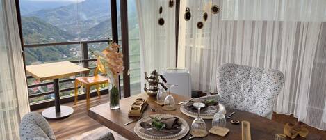 A dining area with a table set for two, a vase with flowers, and a view of mountains through large windows.
