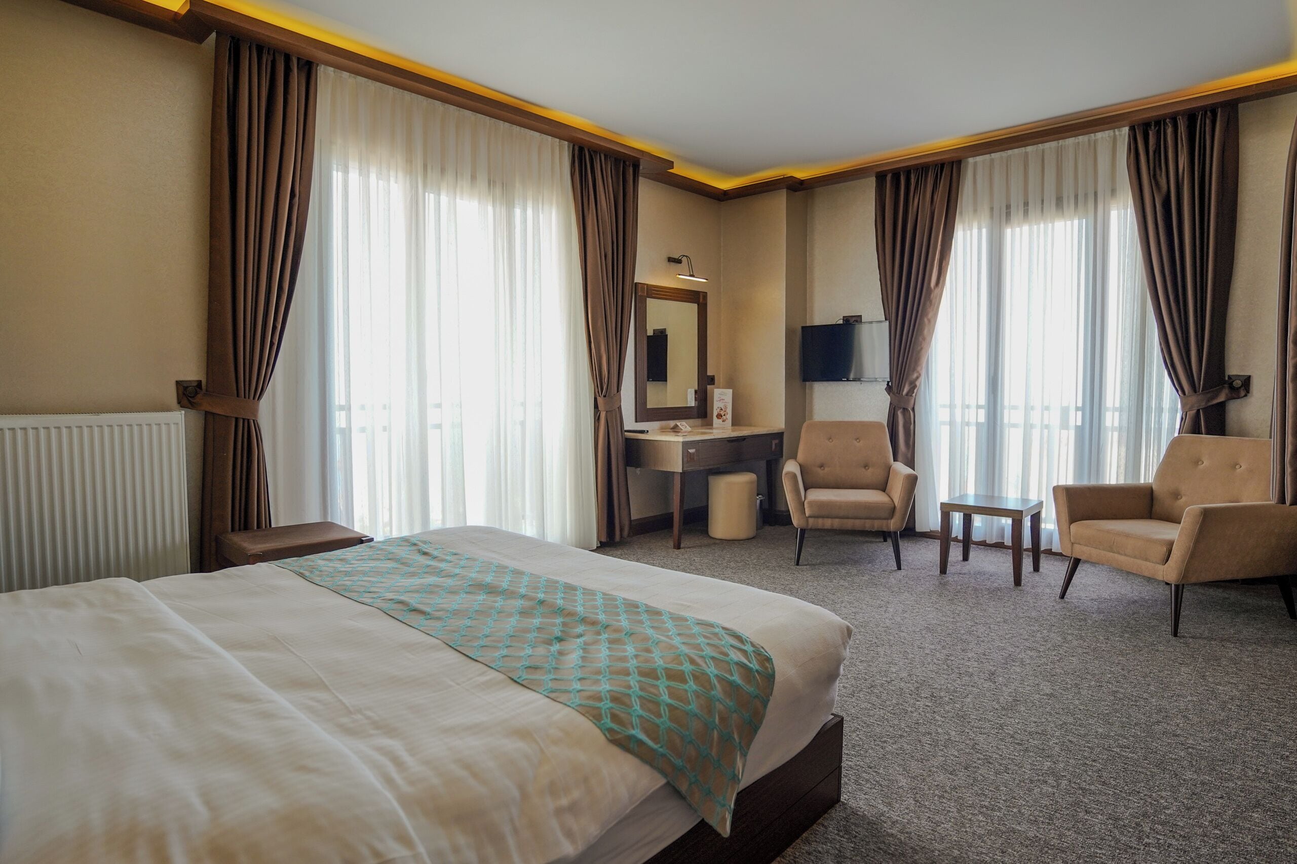 Una habitación de hotel con una cama, un escritorio, dos sillas, un televisor y ventanas grandes con cortinas.