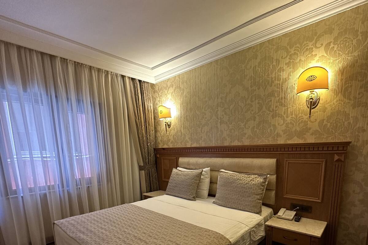 Une chambre d’hôtel avec un grand lit, deux tables de chevet, une petite table, une lampe et une fenêtre avec des rideaux.