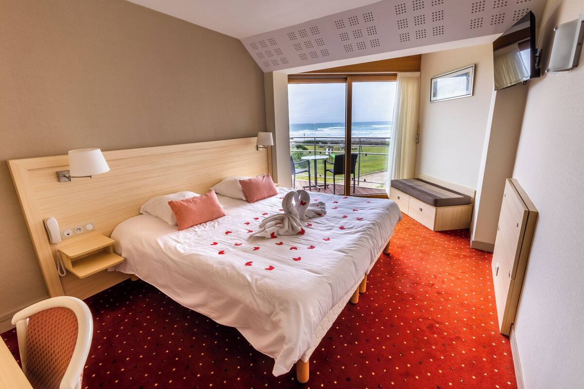 Habitación de hotel con una cama grande, un escritorio pequeño, una silla, un televisor y un balcón con vista al mar.