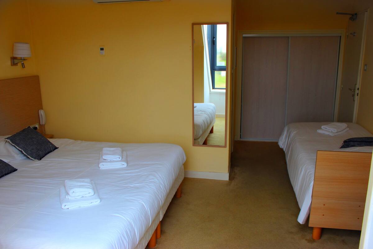 Habitación de hotel con dos camas, cada una con toallas ordenadas, una lámpara de noche y un espejo que refleja la habitación.