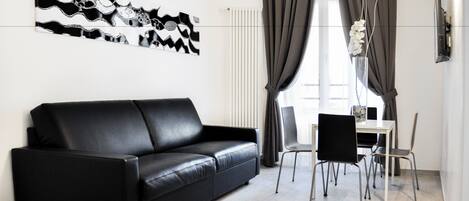 A modern living room with a black leather sofa, a dining table with chairs, and a large abstract art piece on the wall.
