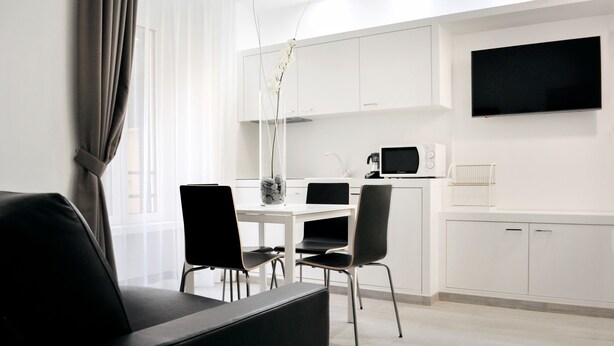 Un salon moderne avec un canapé noir, une table à manger avec des chaises, un coin cuisine avec des meubles blancs, un téléviseur à écran plat et un vase rempli de fleurs.