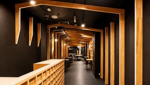 A modern interior with a central wooden reception desk, multiple tables, and chairs arranged along the walls. - Namba Guesthouse HIVE - Hostel (Osaka)