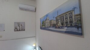 Una habitación con un cuadro grande de un edificio histórico, una pequeña estantería con adornos y un aire acondicionado de pared.