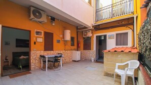 Un pequeño patio al aire libre con una mesa y sillas, una silla de plástico blanco, un muro de piedra y un balcón con techo rojo.