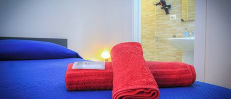 A neatly made bed with blue bedding and two red towels, a bedside lamp, and a bathroom with a sink and mirror.