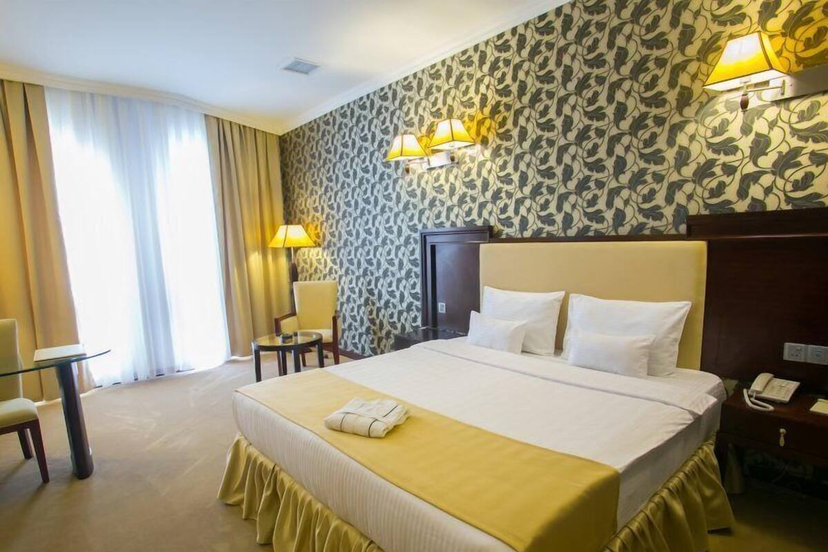 A hotel room with a large bed, a desk, a chair, a telephone, and a window with curtains.