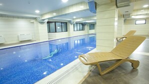 En indendørs swimmingpool med klare blå fliser, en liggestol og en flisebeklædt væg med vinduer.