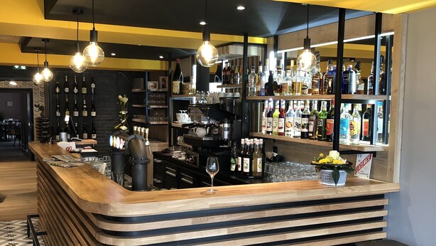 A bar area with a wooden counter, multiple stools, a wine glass, and shelves stocked with bottles. - Le Chêne Vert (Savenay)