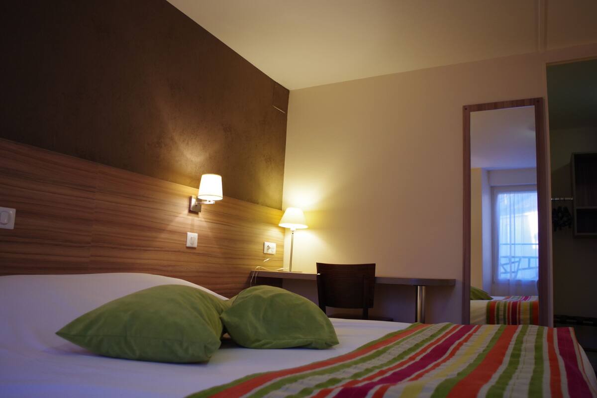 A hotel room with a bed, two green pillows, a striped blanket, a desk with a chair, and a lamp.