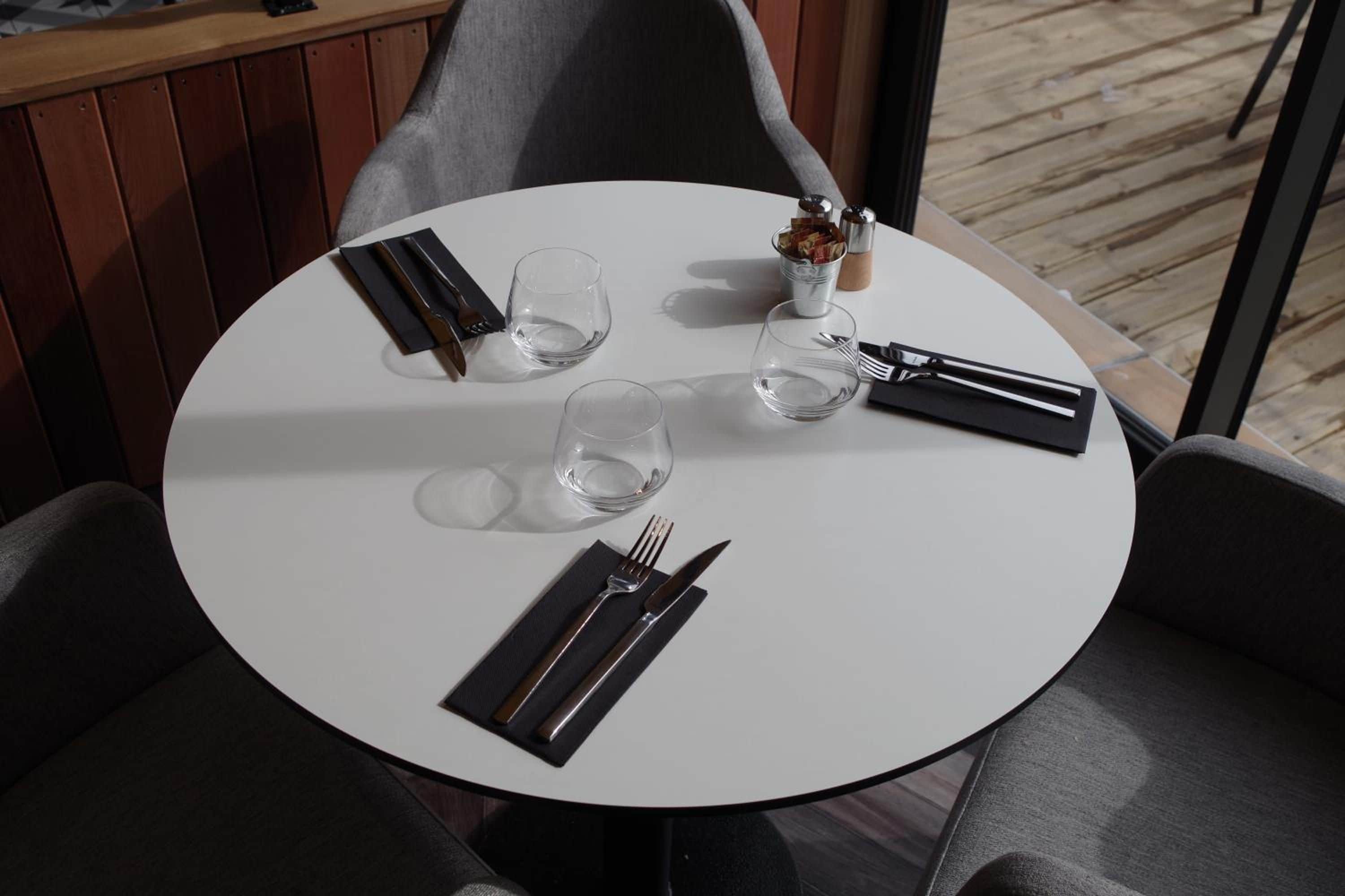A round dining table with a white tablecloth, two empty glasses, a small vase with flowers, and neatly arranged cutlery.