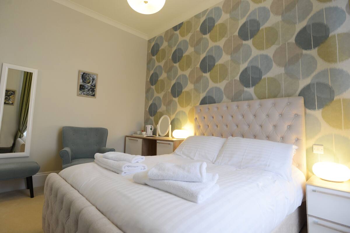 A neatly made bed with white linens and towels, a tufted headboard, a bedside table with a lamp, a chair, and a wall with a circular pattern.