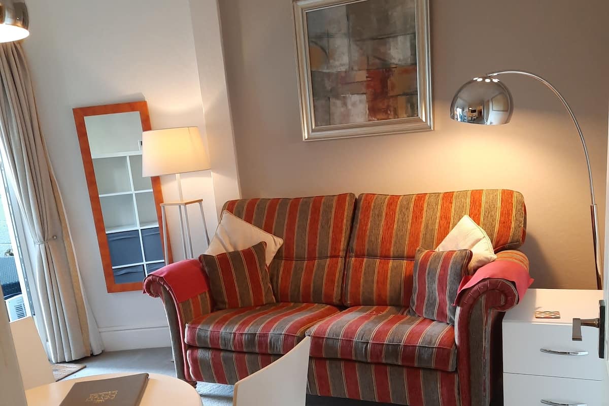 A modern hotel room with a striped sofa, a desk with a laptop, a standing lamp, and a large mirror.