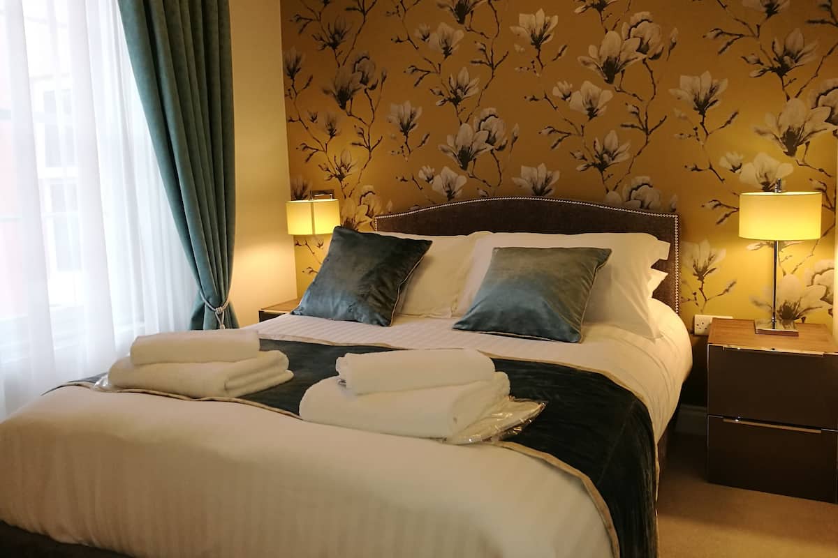 A neatly made bed with pillows and folded towels, a bedside lamp, and a floral wallpaper pattern.