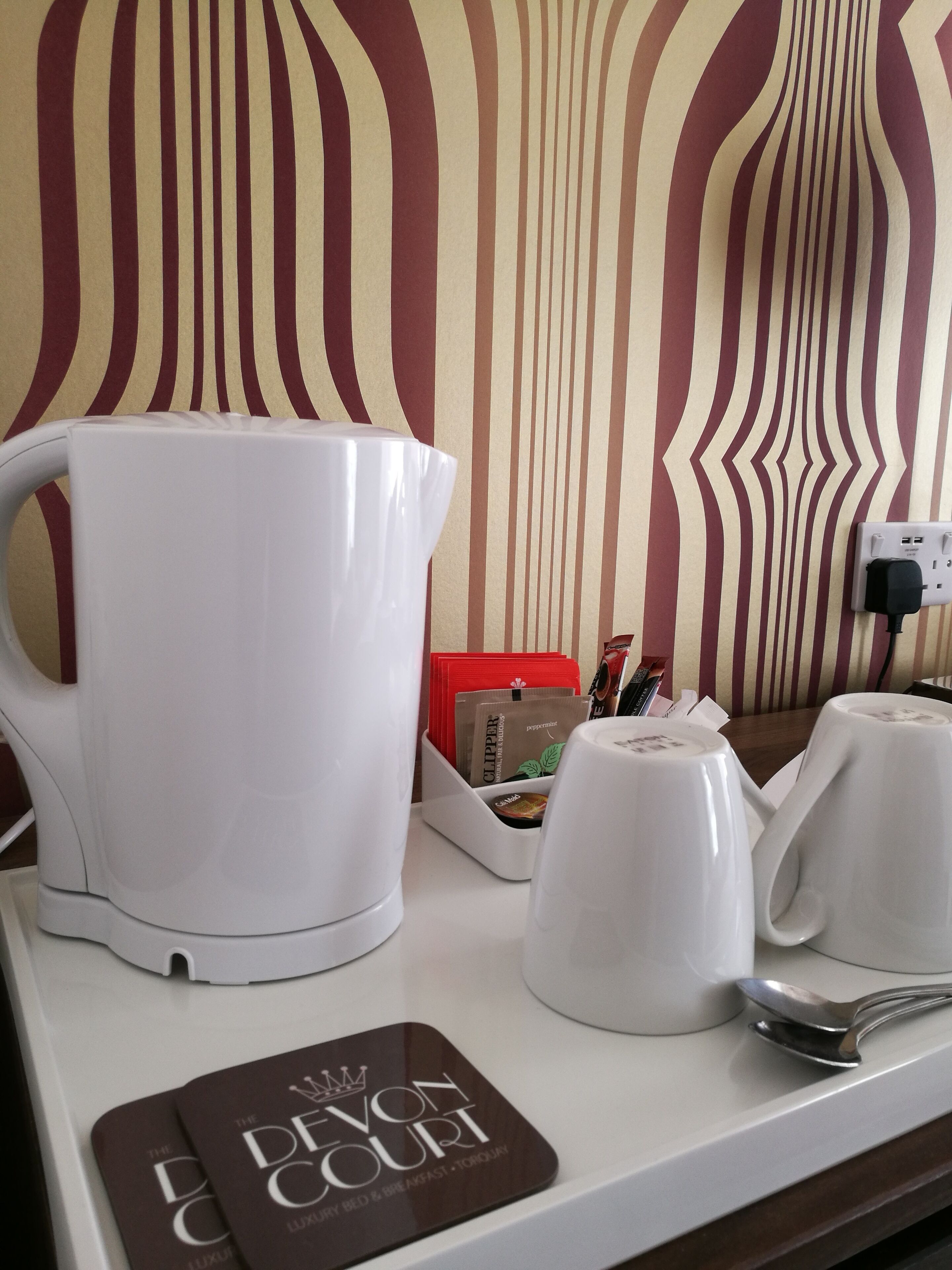 Un plan de travail de cuisine avec une bouilloire blanche, deux tasses blanches, une cuillère et un plateau contenant une dosette de café et un classeur rouge.