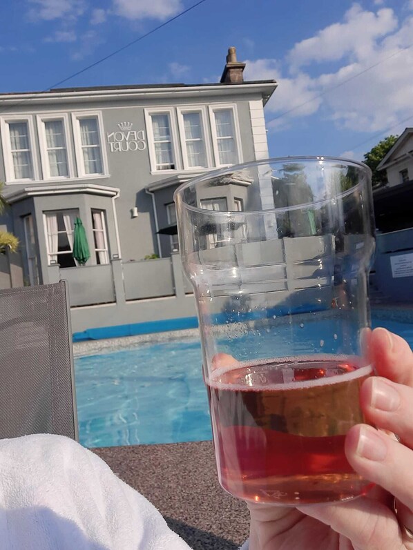 A clear glass with a red liquid, held in front of a pool, with a house in the background. - The Devon Court (Torquay)