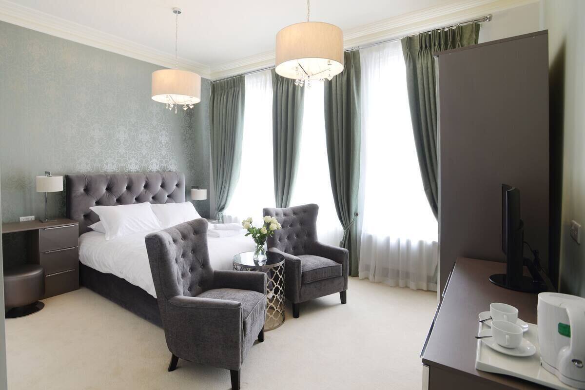 A hotel room with a bed, two armchairs, a small table with a vase, a desk with a television, and a window with curtains.