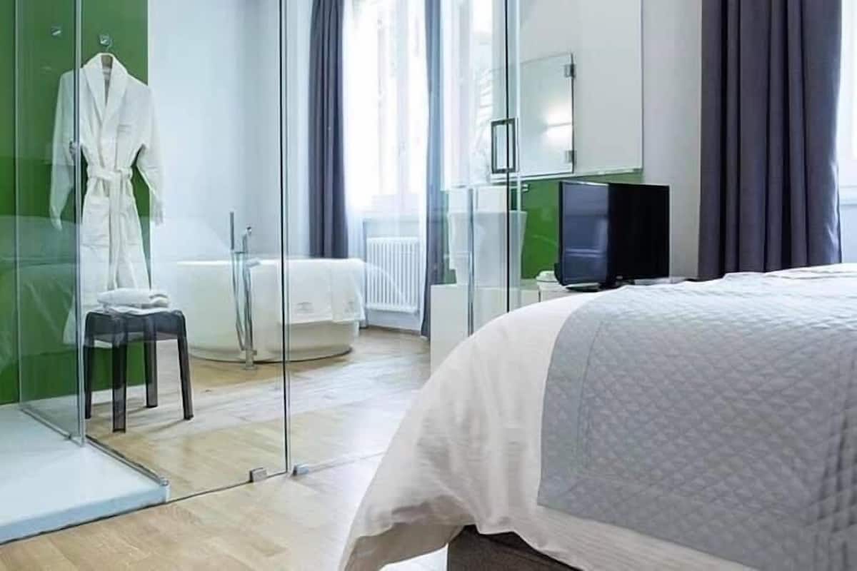 Ein modernes Hotelzimmer mit Duschkabine aus Glas, einer weißen Badewanne, einem Fernseher und einem Bett mit weißer Daunendecke.
