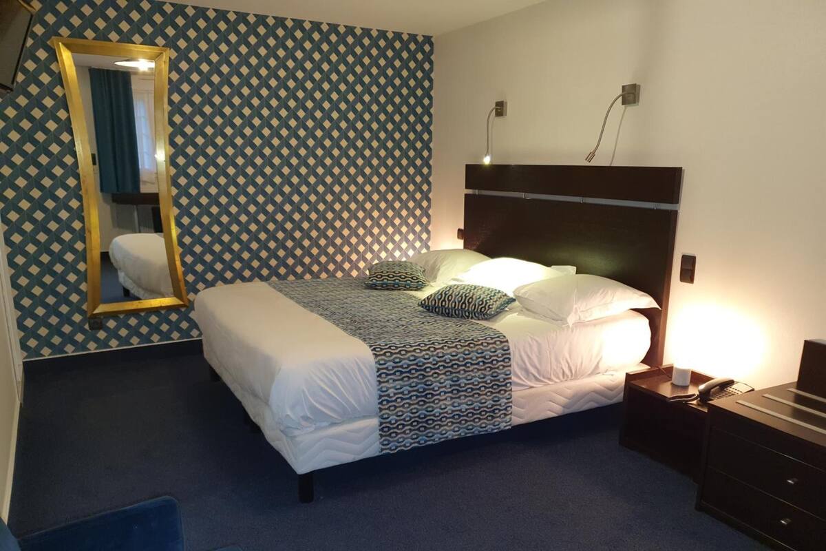Una habitación de hotel con papel tapiz estampado, una cama con ropa de cama blanca y almohadas estampadas, una mesita de noche con una lámpara y un espejo en la pared.