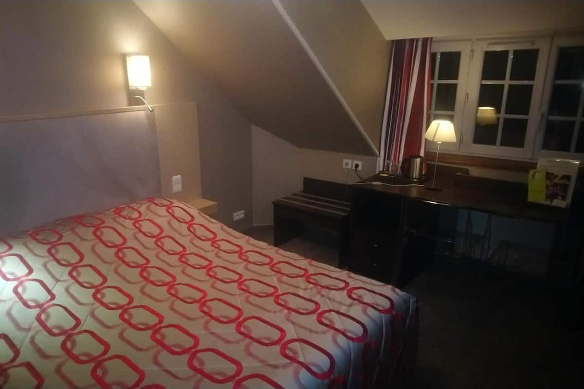 Habitación de hotel con cama, escritorio, silla, lámpara y ventana con cortinas.