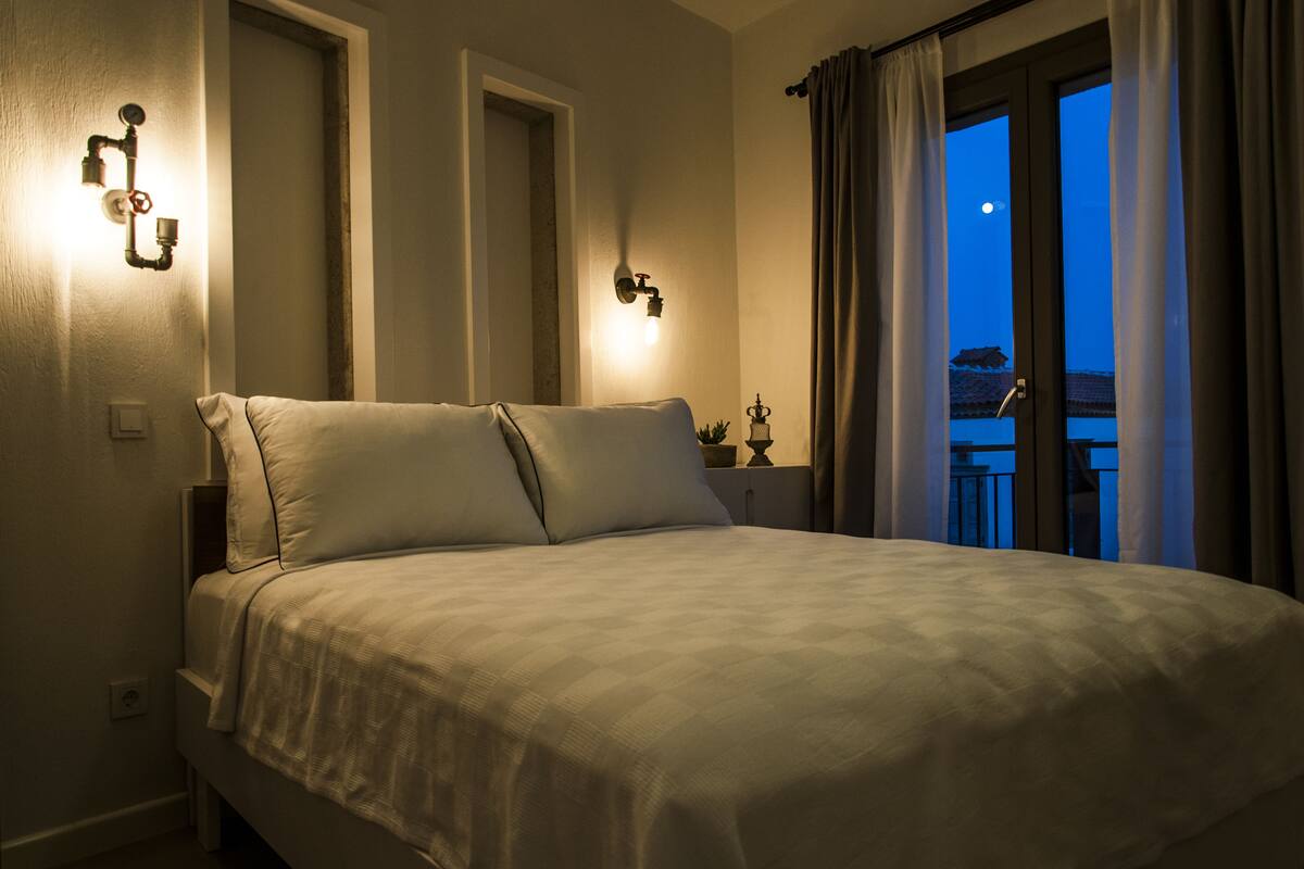 A bedroom with a bed, two wall-mounted light fixtures, a nightstand with a lamp, and a view of a balcony with curtains.