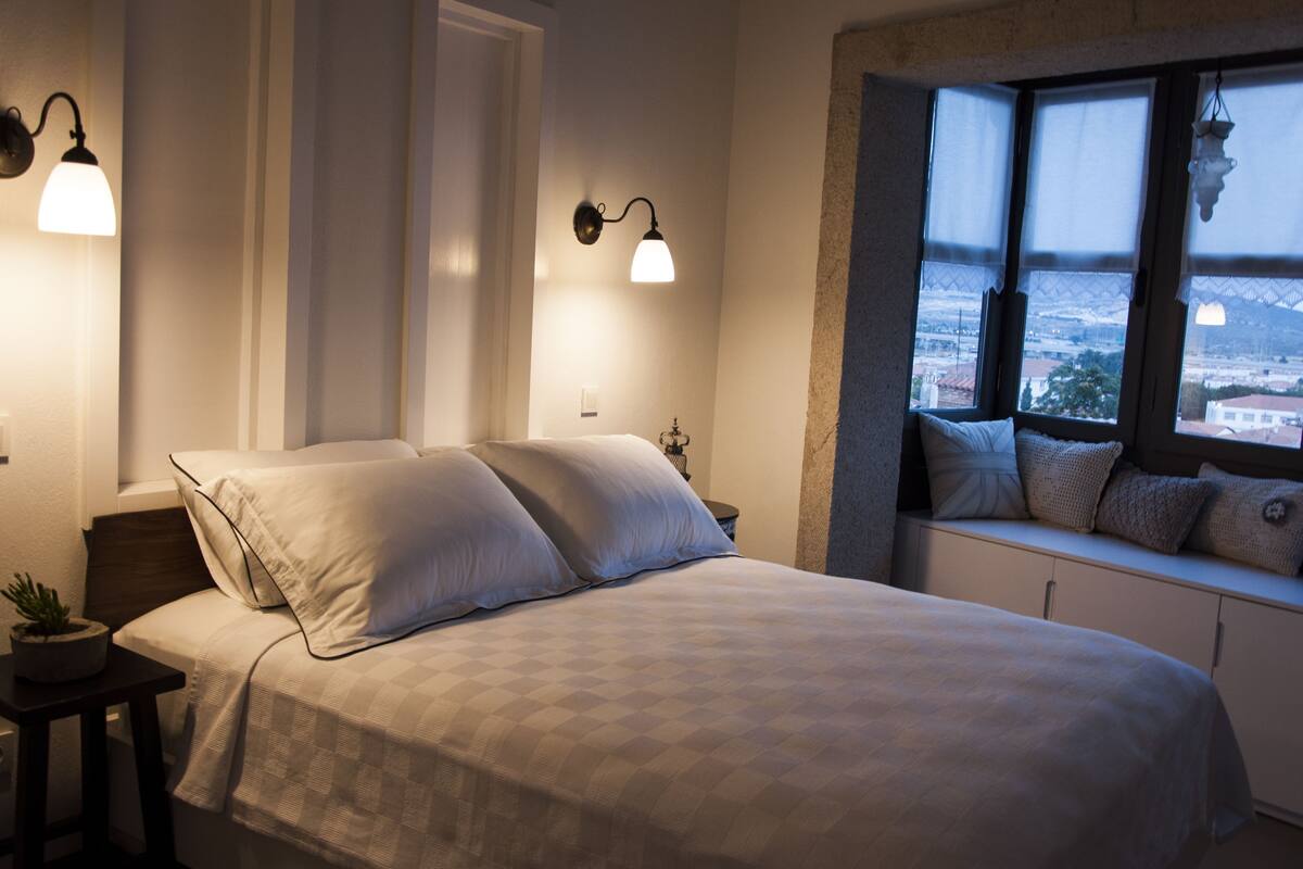 A bedroom with a bed, bedside lamps, a window with a view, and a small table with a plant.