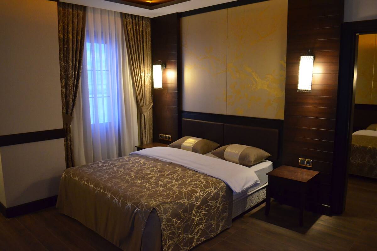 Habitación de hotel con una cama grande, una mesita de noche, una lámpara de pared y una ventana con cortinas.