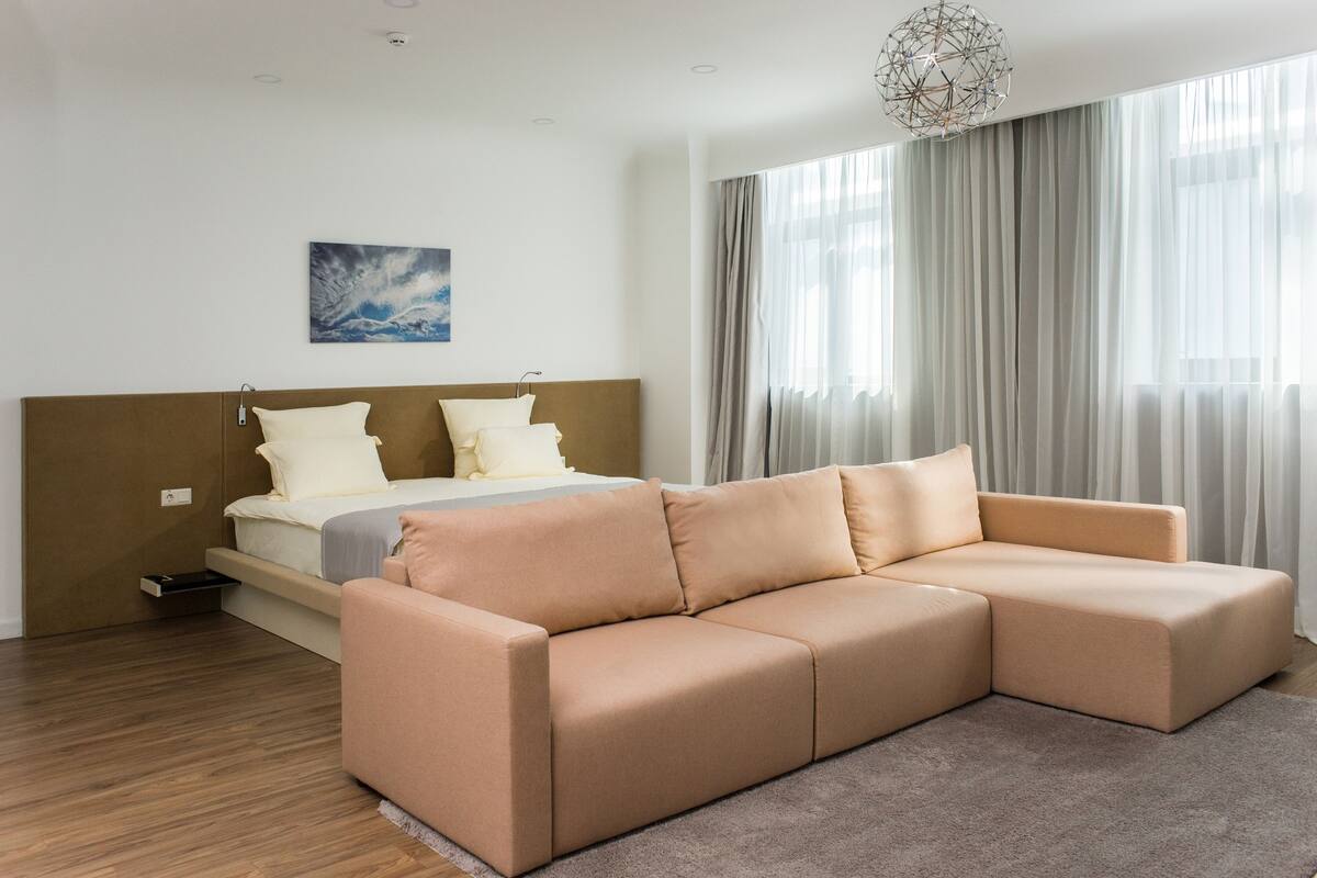 A modern bedroom with a large bed, a beige sectional sofa, a bedside table, and a large window with sheer curtains.