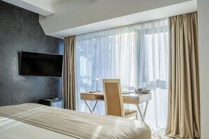 A hotel room with a bed, a desk with a chair, a flat-screen TV, and a window with sheer curtains. - Marlin Hotel (Odesa)