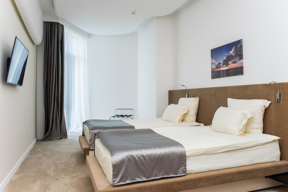 A modern hotel room with a large bed, a bedside table, a flat-screen TV, and a view of the sea through the window.