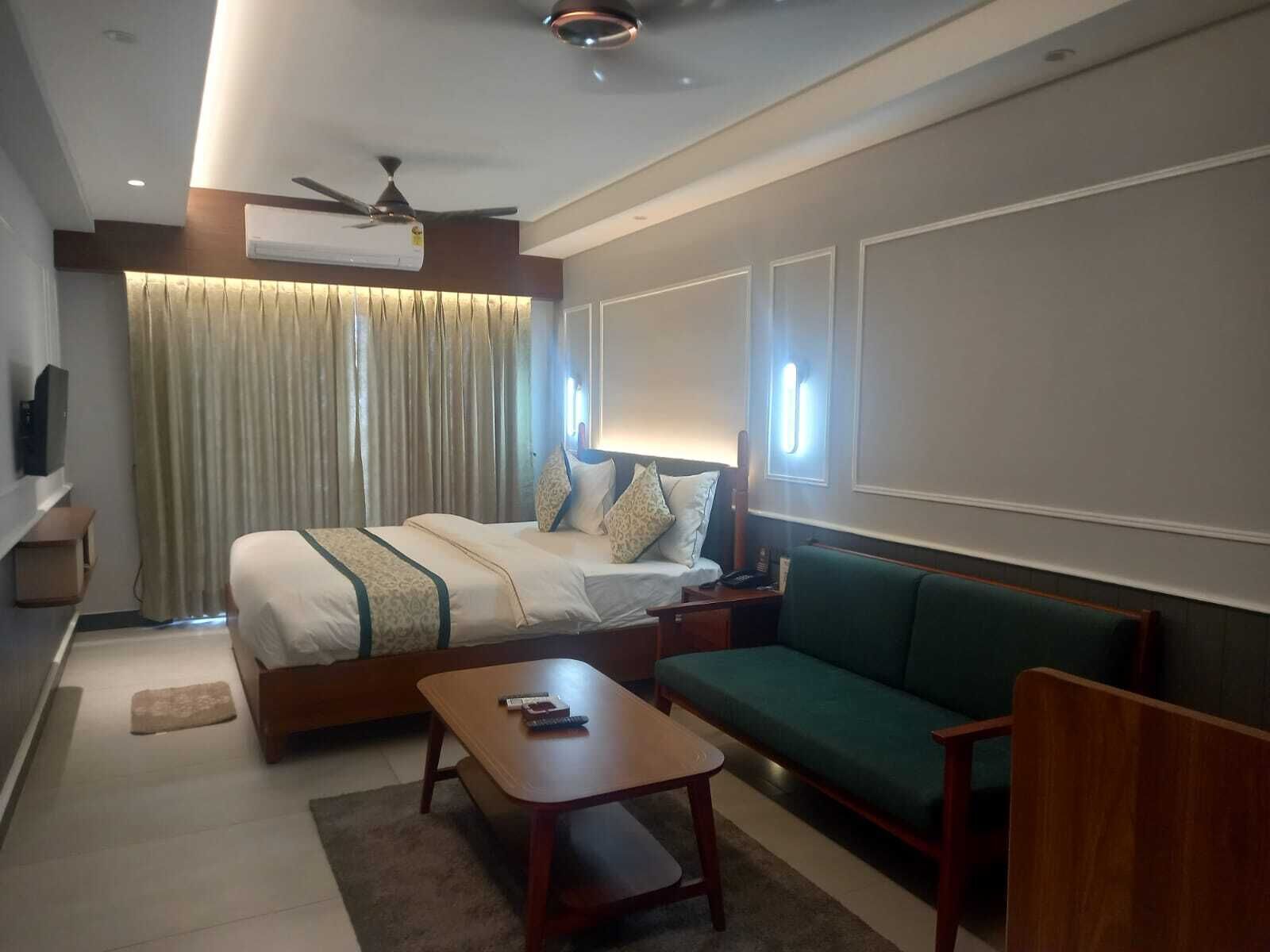 A modern hotel room with a large bed, a green sofa, a wooden coffee table, and a ceiling fan.