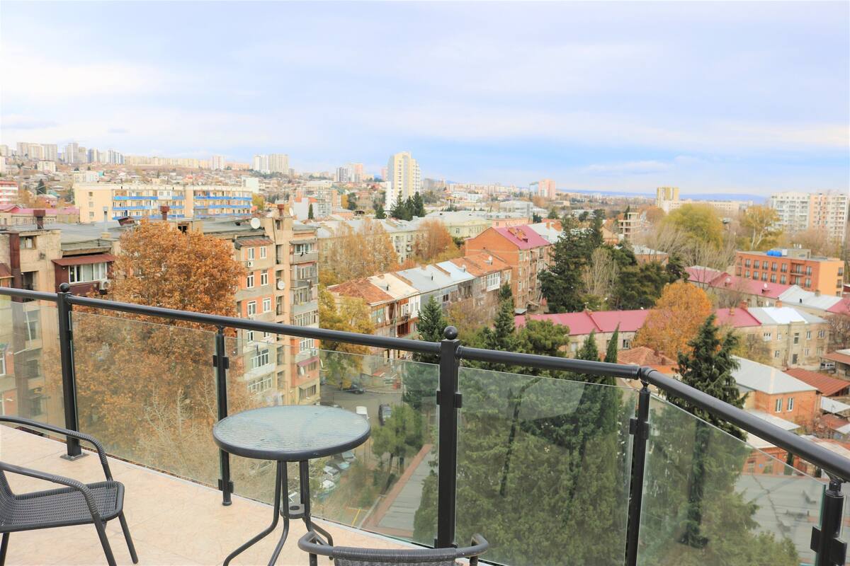 A balcony with a glass railing overlooking a cityscape with various buildings and trees.