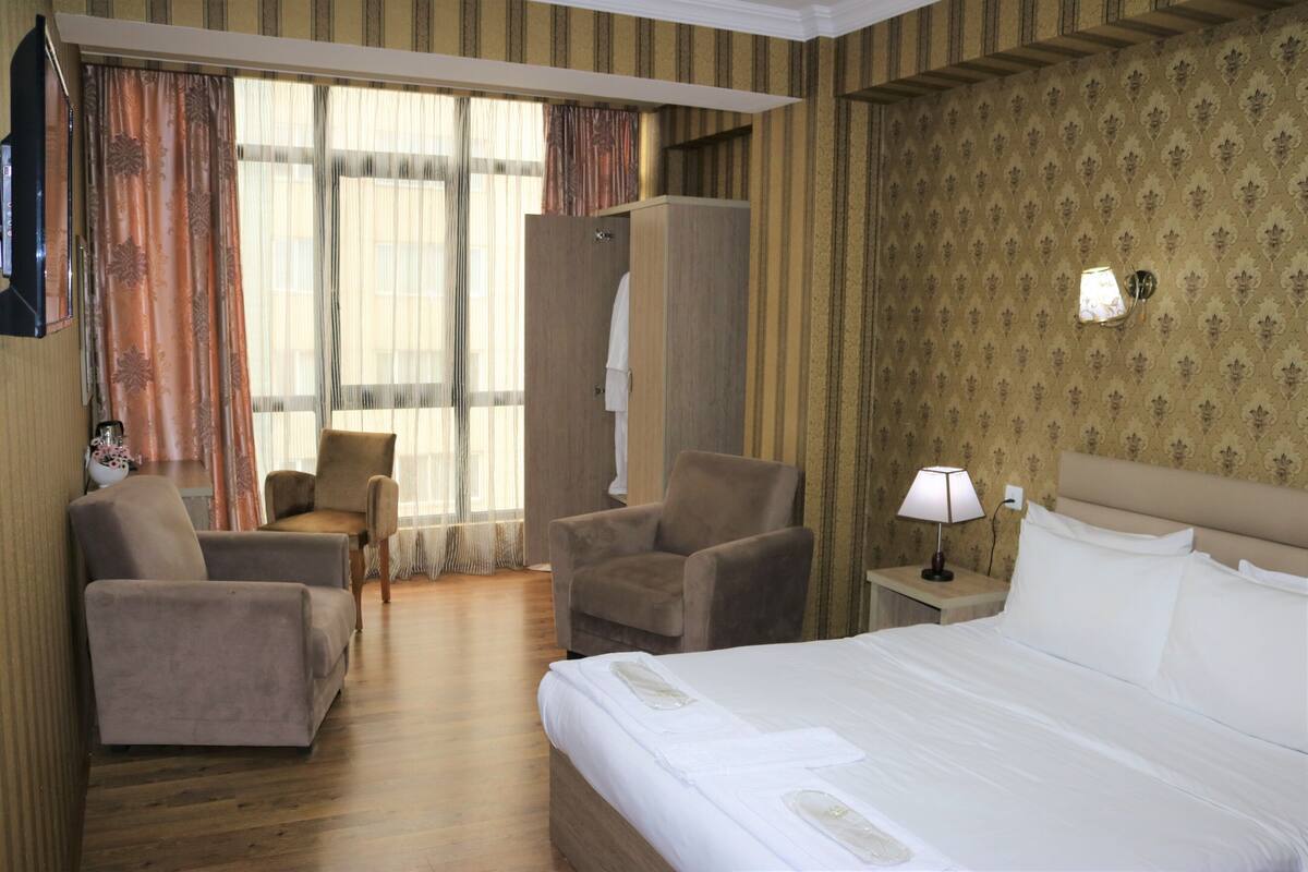 A hotel room with a large bed, two armchairs, a sofa, a wardrobe, and a nightstand with a lamp.