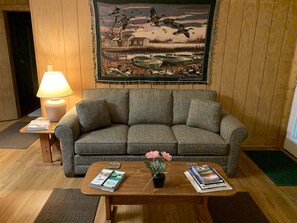 A cozy living room with a grey sofa, a wooden coffee table with magazines and a potted plant, a lamp, and a framed painting of a duck pond. - Misty Creek Log Cabins (Maggie Valley)