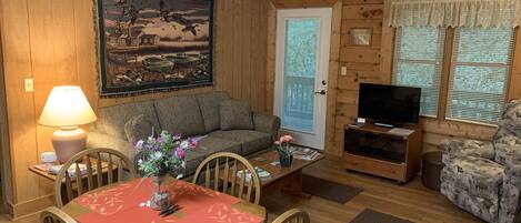 Ein gemütliches Wohnzimmer mit Holzboden, einer Couch, einem Couchtisch mit Blumenvase, einem Esstisch mit Stühlen und einem Fernseher auf einem Schrank.